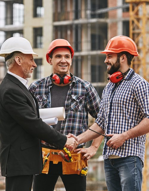 Good job! Cheerful elderly engineer in formal wear and white helmet holding engineering drawings and shaking hands of two happy young builders while standing at construction site. Architectural project. Construction concept. Building concept